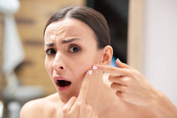 Expressive young woman feeling disappointed and scared while showing a pimple on her cheek