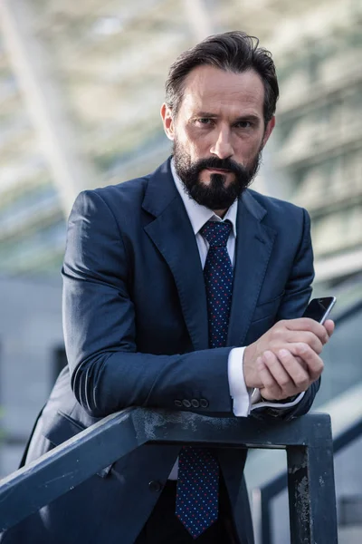 Serious attitude. Pleasant serious businessman holding a phone while looking at you