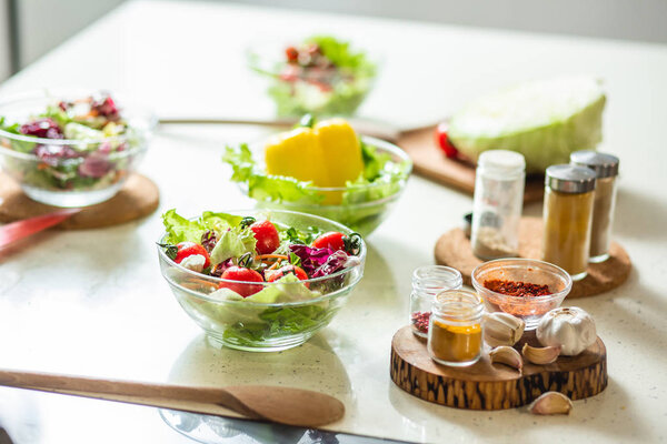 Close up of glass bowls with fresh vegetable salads and spices in glass bottles standing next to them on the pieces of wood