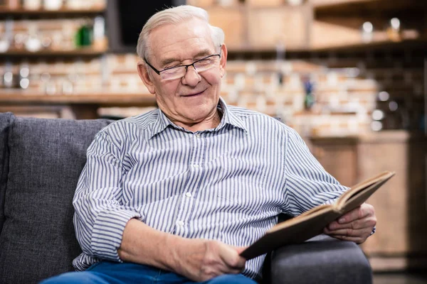 Eternal wisdom. Positive aged man reading a book while expressing happiness at home