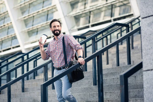 Get some energy. Positive adult man drinking coffee while walking down the stairs