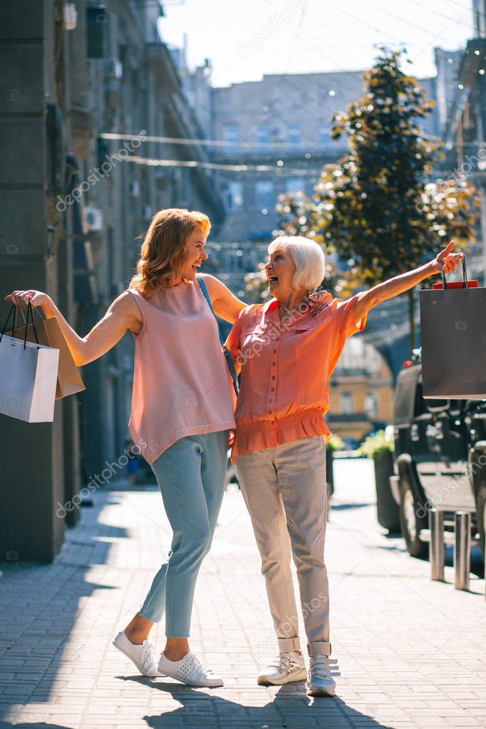 Two mirthful women smiling to each other in the street with shopping bags in hands