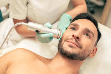 Yanağına yakın oksijen meso terapi aracı tutan lastik eldiven genç neşeli bir adam ve güzellik yüz