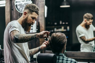 Professional barber with tattooed arms holding a comb and making haircut for his client