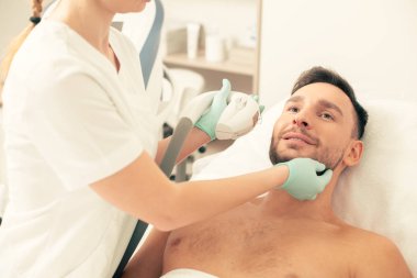 Sakin sakallı adamın yanında duran ve fotoğraf gençleştirme işlemi sırasında yüzüne dokunan tıbbi üniformalı profesyonel kozmetolojist