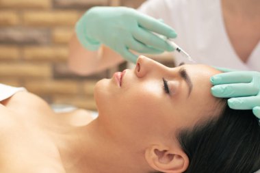 Gözleri kapalı yatan genç kadının yan görünümü ve hyaluronik asit enjeksiyonu yapan kozmetolojist