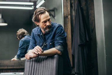 Düşünceli orta yaşlı sakallı adam kol sandalyeye ellerini koyarak ve berber dükkanı nda iken başını çevirerek