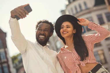 Neşeli genç adam ve kadın rahat yazlık giysiler içinde açık havada duran ve selfie çekerken akıllı telefon kameraya gülümseyerek