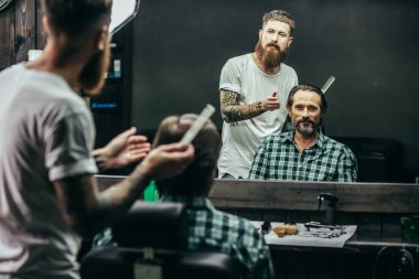 Calm bearded barber standing and thinking while his satisfied client sitting in the armchair in front of the mirror and smiling