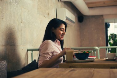 Koyu saçlı genç bayan bir kafede ahşap masada oturan ve onu salata tadını çıkarırken gülümseyerek