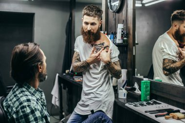 Genç sakallı berber meraklı müşterisinin önünde duruyor ve omzunda büyük bir dövme gösteriyor.