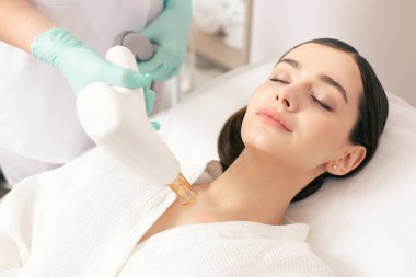 Koyu saçlı bayan kapalı gözlerle yalan süre profesyonel bir sağlık çalışanı modern bir araç tutarak ve bir lazer ile izleri kaldırma