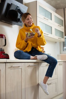 Mutlu sarı kazak genç bayan modern mutfakta tezgahüzerinde rahatça oturan ve salata yerken bir gülümseme ile mesafe içine bakarak