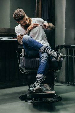 Sakin konsantre genç berber dövmeli kolları ile başını eğik ve dikkatle makas jilet bakarak