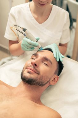 Kapalı gözlerle yatan gömleksiz adam ve alnındaki deriyi besleyen dikkatli güzellik uzmanı