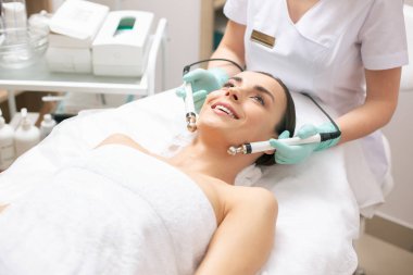 Klinikte mikrocurrent tedavi görürken tıbbi kanepede yatan ve heyecanlı görünen mutlu genç bayan