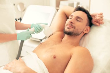 Genç adam kapalı gözlerle tıbbi kanepede yatan ve koltuk altında lazer epilasyon prosedürü geçerken gülümseyerek