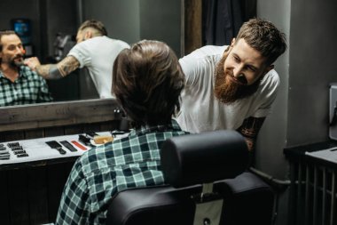 Emotional bearded barber smiling and leaning to the client while combing his beard