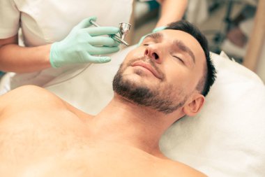 Kapalı gözlerle yatan ve cildine oksijen spreyi tedavisi gören sakin sakallı bir adamın yakın çekim