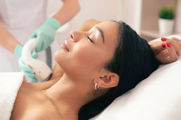 Young beautiful woman lying with her eyes closed and smiling while undergoing laser hair removal on her armpit