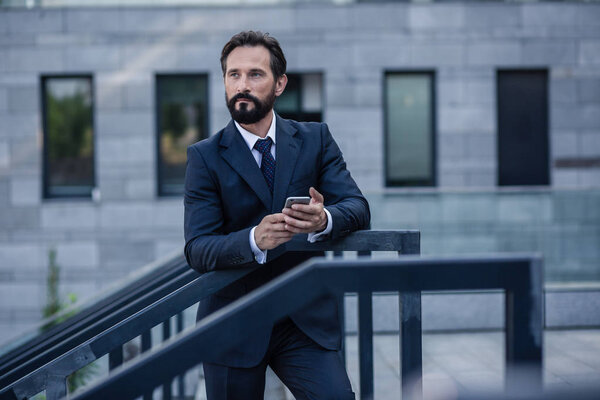 Stay confident. Serious bearded businessman leaning on the handrail while using his smartphone