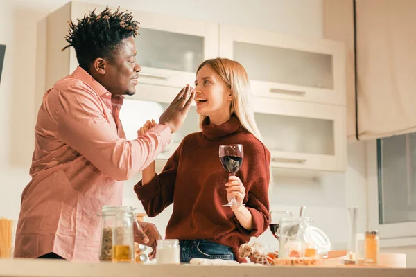 欢快的年轻女士站在厨房里 喝了一杯酒 而她的男朋友在她的鼻子上放了一些面粉 — 图库照片