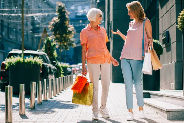 Positive expressive ladies of different ages standing in the street with shopping bags and smiling to each other