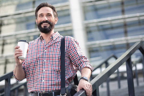 Open minded person. Cheerful bearded man walking in the city while drinking coffee