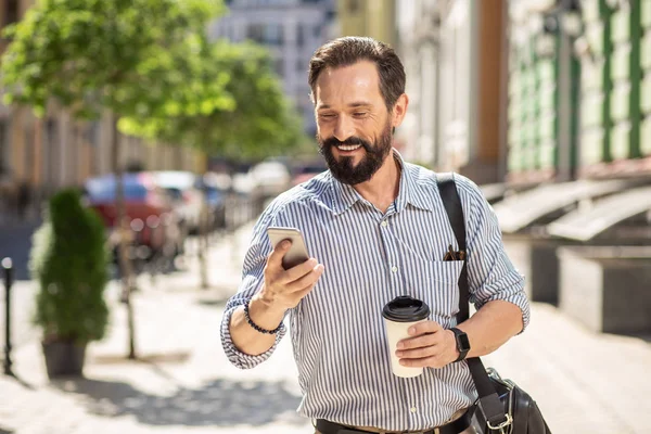 Hızlı yaşam. Pozitif gülümseyen yetişkin adam sokakta giderken akıllı telefon kullanarak