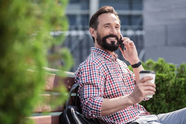 Small talk. Waist up of a cheerful bearded man drinking coffee and talking on phone on the bench