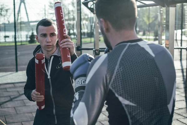 Young professional sports trainer being outdoors with his boxer and putting red boxing sticks up for him