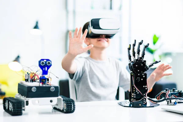 Selective focus of robotic devices standing on the table while ingenious young boy testing them with VR device
