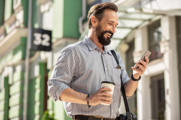 On my way to work. Waist up of cheerful bearded man using his smartphone and drinking coffee