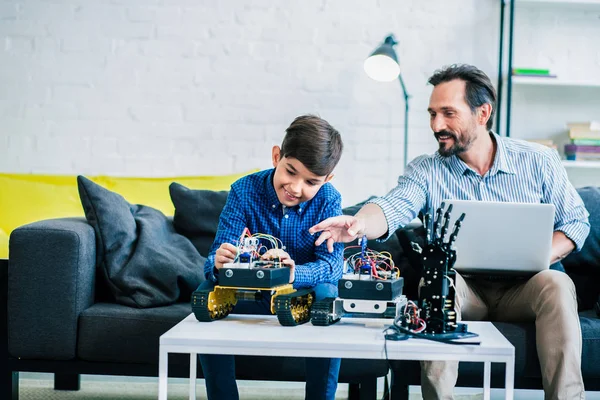 快乐的成年男子使用他的笔记本电脑 同时帮助他的儿子与机器人技术在家里 — 图库照片