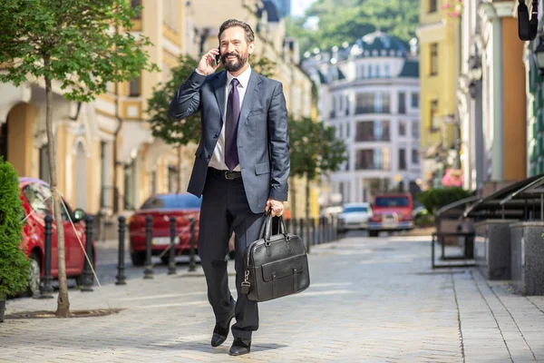 Nice talk. Cheerful smiling businessman walking to work while talking on phone