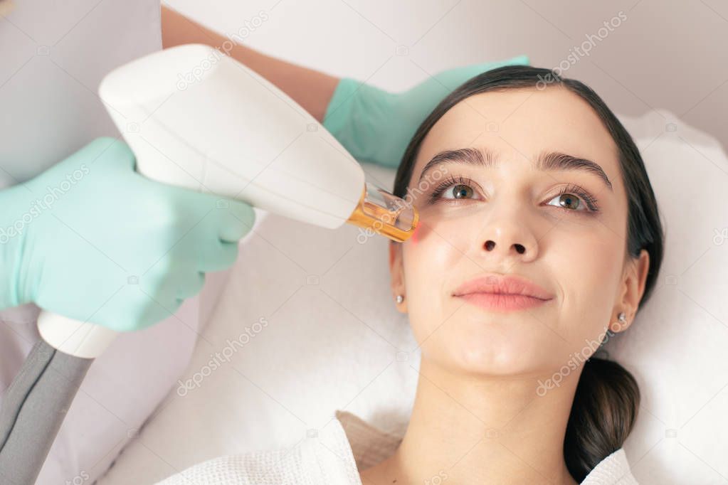 Beautiful lady lying and smiling while hand in rubber glove holding a modern tool and removing stretch marks on her face