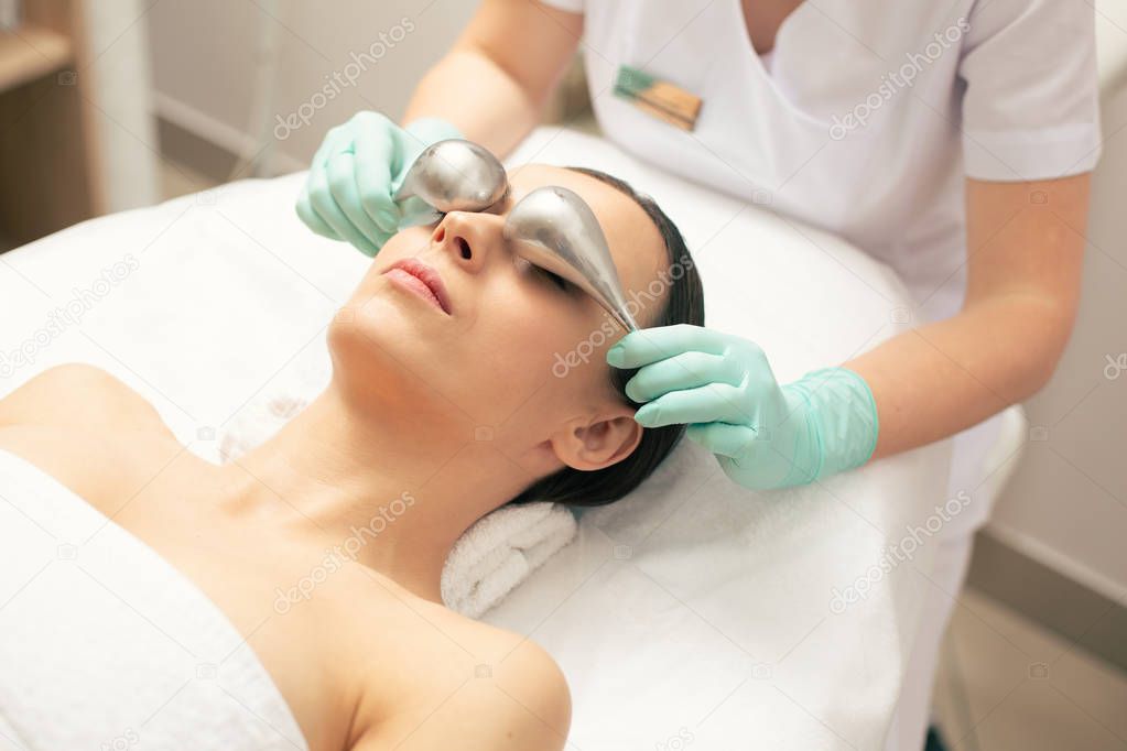 Calm young woman lying on the medical couch and professional cosmetologist using cryo-sticks at the procedure