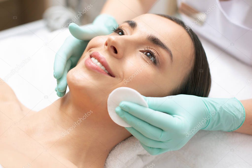 Close up photo of the relaxed woman lying on the towel and smiling while hand in rubber glove cleaning her face with a cotton pad