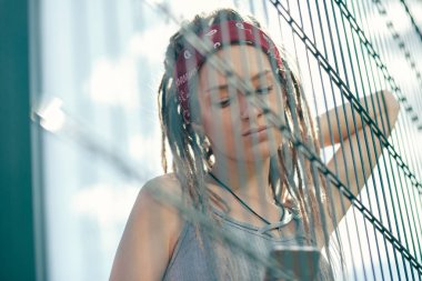 Dreadlocks zincir bağlantı çit yanında duran ve dikkatle onu modern cihazın ekranında bakarak düşünceli genç kadın