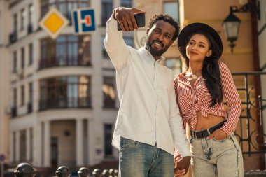 Genç yetişkin çift sokakta el ele tutuşuyor ve birlikte selfie çekerken gülümsüyor