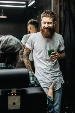 Uzun sakallı duygusal neşeli berber, ziyaretçisinin önünde durup dinlerken ona gülümsüyor.