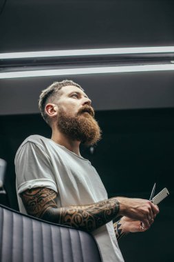 Kollarında makas ve tarak tutan ve mesafe içine bakarak dövmeler ile Profesyonel sakallı adam