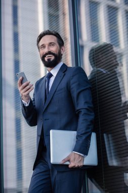 Profesyonel tavır. Handsoem duvara yaslanmış bir dizüstü bilgisayar ve akıllı telefon tutan gülümseyen işadamı