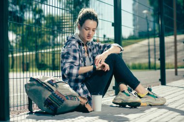 Sakin genç kadın rahat giysiler giyen ve zincir bağlantı çit yanında otururken onu saat bakarak