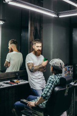 Sakallı profesyonel berber berber dükkanı tezgahın yanında duran ve onu dinlerken müşterisine bakıyor