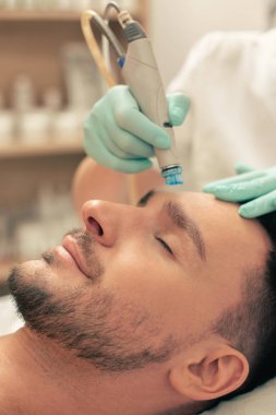 Sakallı adam kapalı gözlerle dinlenirken, profesyonel bir kozmetolojist alnının derisini besler.