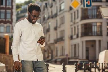 Beyaz gömlekli genç Afro-Amerikan adam sokakta ayakta ve onun gadget bakarken gülümseyerek memnun