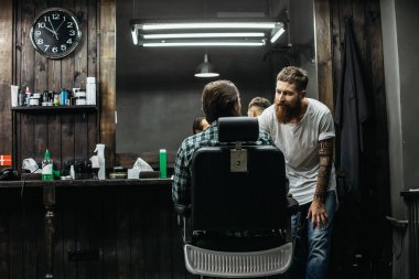 Calm professional long bearded barber standing next to the mirror and bending his back while leaning to the client