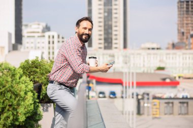 Güzel hava. Çatıda dururken akıllı telefonlarını kullanan neşeli handsoem işadamı