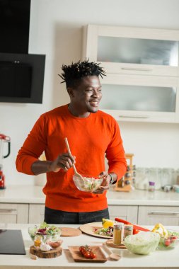 Pozitif genç adam gülümseyerek ve cam kase taze salata karıştırırken mesafe içine bakarak